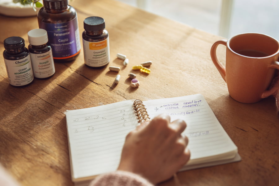Mesa con múltiples frascos de suplementos desordenados y cuaderno con anotaciones de vitaminas, taza terracota, confusión suplementación menopausia